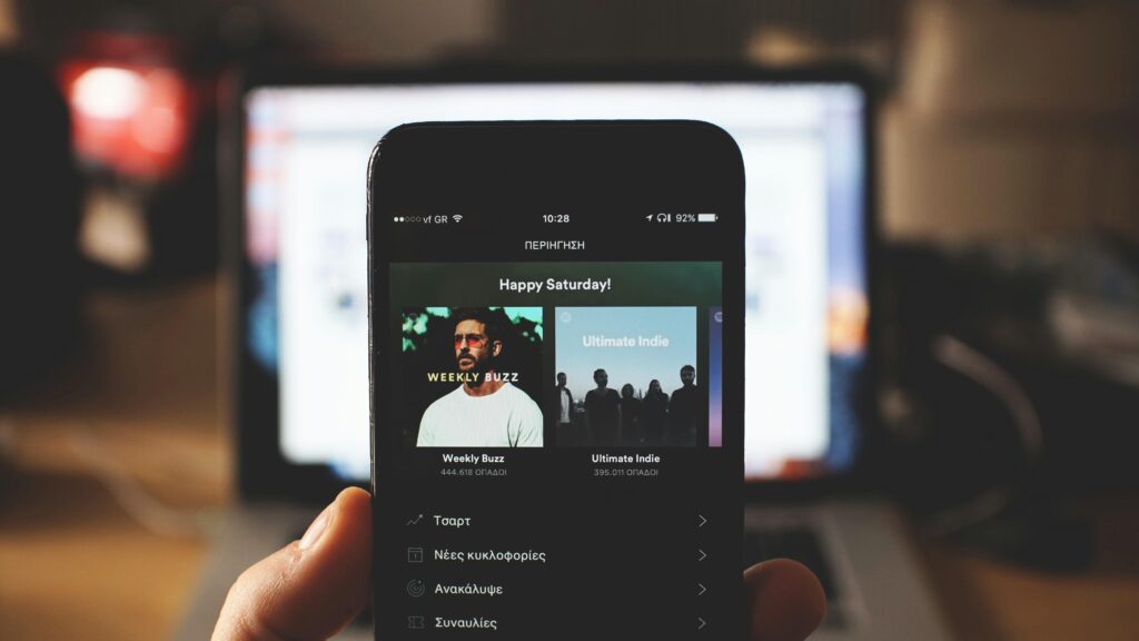 A person holding a smartphone with music streaming app in front of a laptop screen indoors.